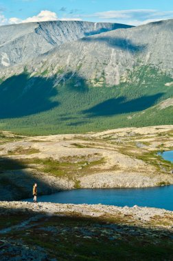 Man in mountain standing near lake and looking at camera from far away clipart