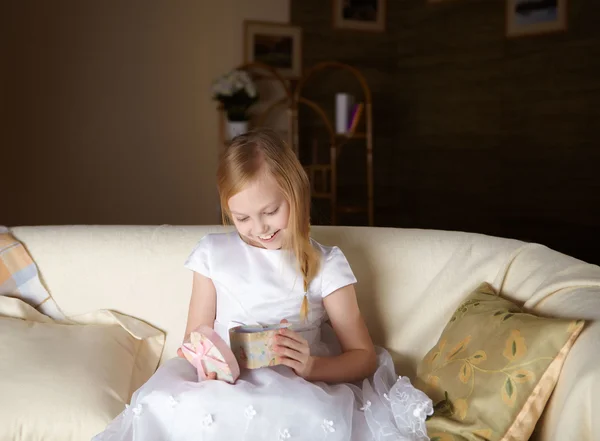 在白色的裙子 坐在一张沙发 一份礼物在家里上漂亮的小女孩 — 图库照片
