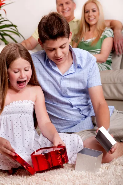 Kids opening christmas gifts with parents in the background — Stock Photo, Image