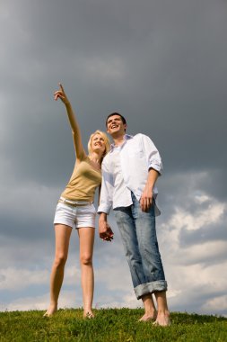 Happy young couple looking at something above a green meadow. Man show something by hand clipart