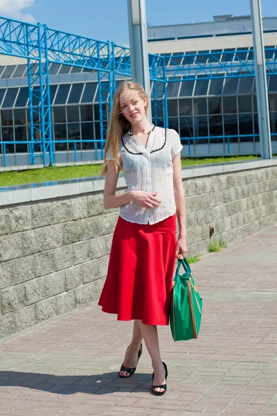 Smiling sexy blonde girl wearing red skirt with a green bag. In — Stock Photo, Image