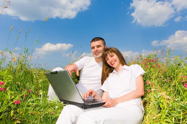 若いカップルのノート パソコンで芝生のフィールド上に座っています 青い空を背景に — ストック写真