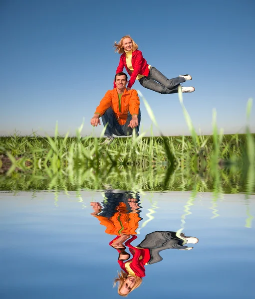 Happy smiling couple jumping in blue sky. Reflected in Water. — Stock Photo, Image