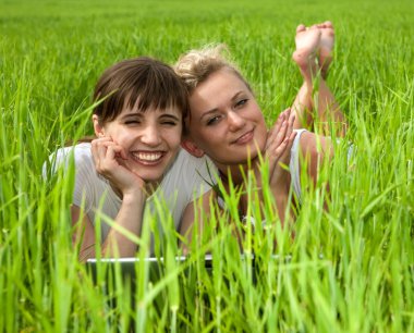 Two beautiful girls in white clothes are laughing at laptop computer outdoors. Lay on the green grass. clipart