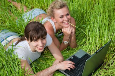 iki güzel kız Beyaz giysili gülüyor ve açık havada dizüstü bilgisayar arıyor. Yeşil çimenlerin üzerinde yatıyordu.