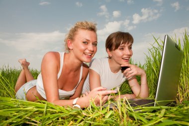 iki güzel kız Beyaz giysili gülüyor ve açık havada dizüstü bilgisayar arıyor. Yeşil çimenlerin üzerinde yatıyordu.