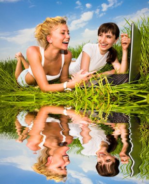 Two beautiful girl in white clothes on a laptop computer outdoors. Lay on the green grass. clipart