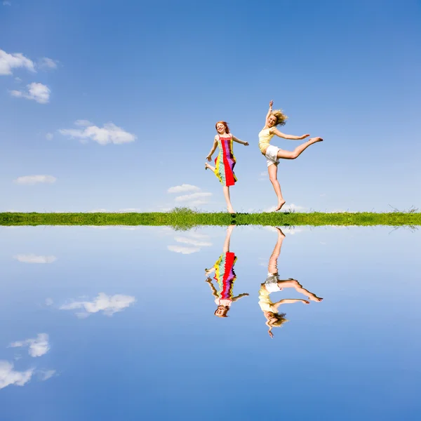 Dos chicas felices saltando juntas en el prado verde. Reflejado en wa —  Fotos de Stock