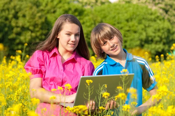 Two Smiling teenagers with laptop on meadow full of yellow flowe — Stock Photo, Image