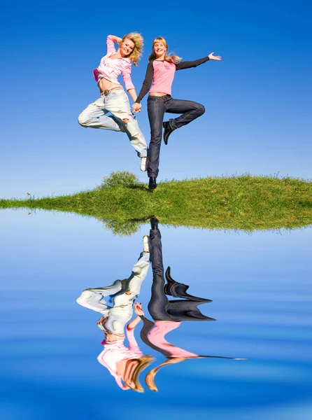 Dos chicas felices saltando en el prado verde. Reflejado en el agua —  Fotos de Stock