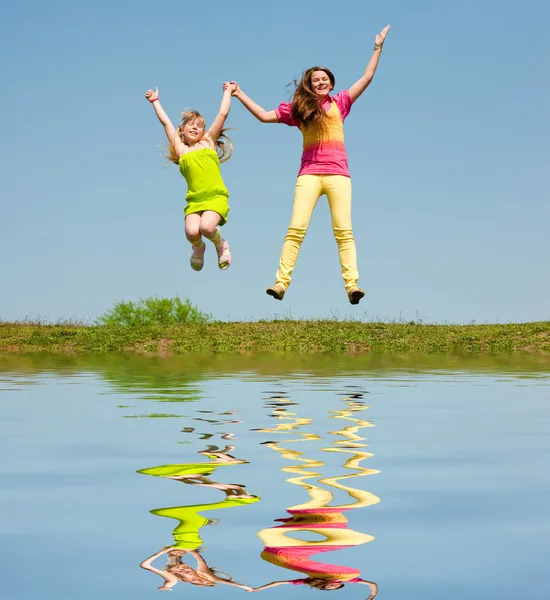 Twee meisjes springen op groene weide. weerspiegeld in water. — Stockfoto