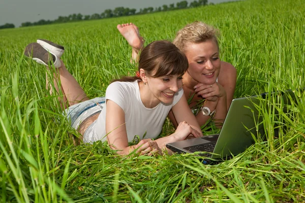 两个漂亮的女孩穿着白色的衣服笑着看着户外的膝上型计算机 躺在绿草地上 免版税图库图片