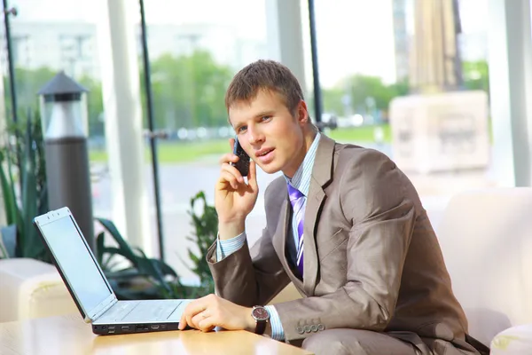 Businessman sitting at table in office hall, talking on mobile phone and us — Stock Photo, Image