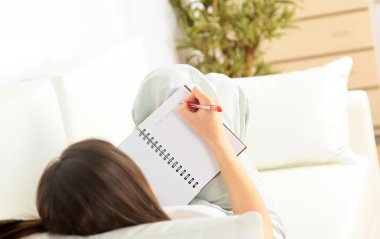 Portrait of a smiling young woman lying on sofa and writing documents clipart