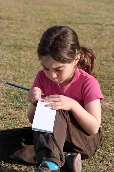 Menina bonito escreve enquanto sentado na grama — Fotografia de Stock