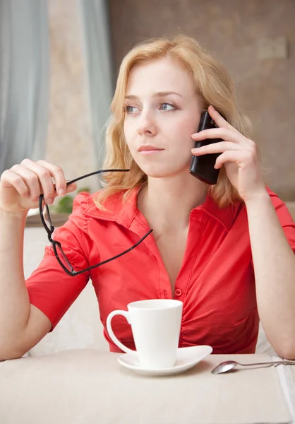 Young blond girl in cafe is drinking coffee and is speaking by mobile phone — Stock Photo, Image