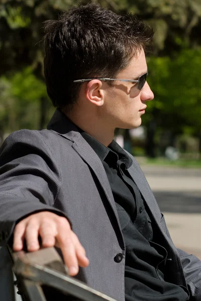 Férfi napszemüveg és kabát ül egy padon közepén — Stock Fotó