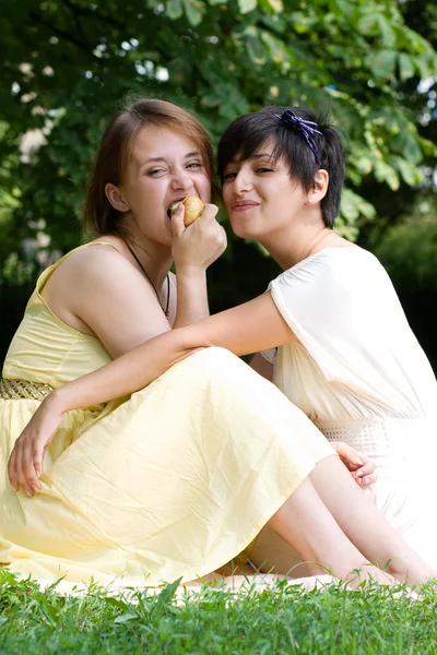 Две девушки едят яблоко на открытом воздухе улыбаясь — стоковое фото