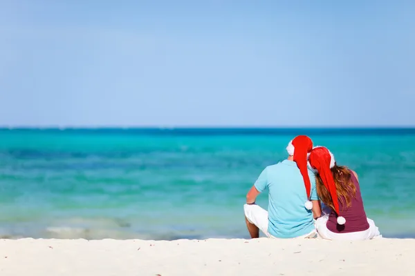 Романтична пара в Санта-капелюхах сидить на тропічному пляжі — стокове фото