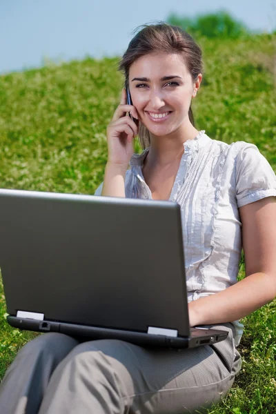 Güzel bir kadın telefonu ve dizüstü bilgisayar ile oturup çimenlerin üzerinde — Stok fotoğraf