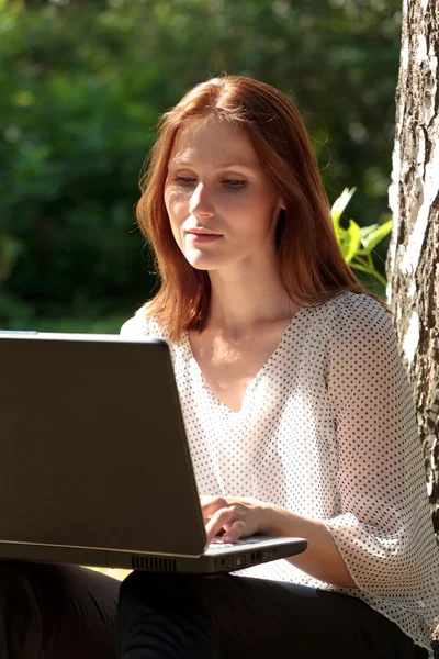 Όμορφη νεαρή γυναίκα που κάθεται σε ένα χόρτο σε ένα πάρκο με το φορητό υπολογιστή. — Φωτογραφία Αρχείου