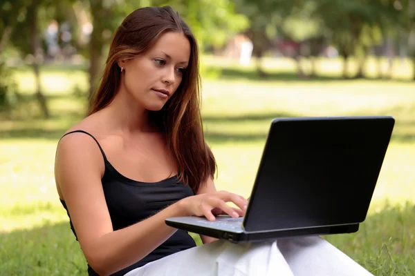 Mooie jonge vrouw zit op een gras in een park met de laptop. Stockfoto