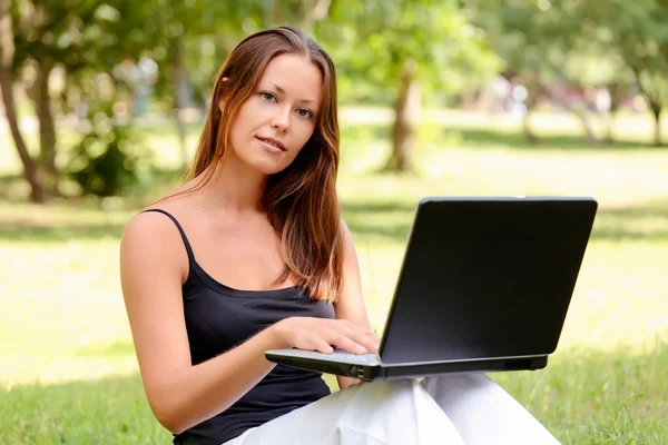 Mooie jonge vrouw zit op een gras in een park met de laptop. — Stockfoto