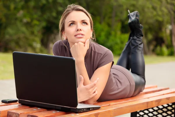 Jovem mulher bonita com laptop deitado no banco em um parque — Fotografia de Stock