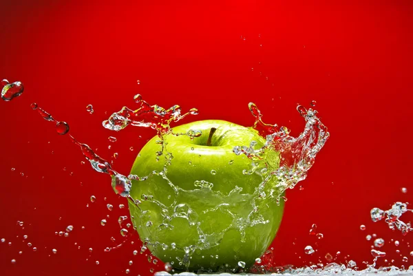 Зеленое яблоко с брызгами воды на красном фоне — стоковое фото