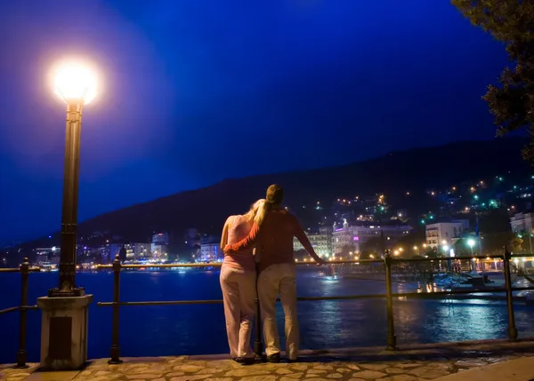 Ζευγάρι στέκεται κοντά στη θάλασσα και κοιτάζοντας το βράδυ πόλη panoram — Φωτογραφία Αρχείου