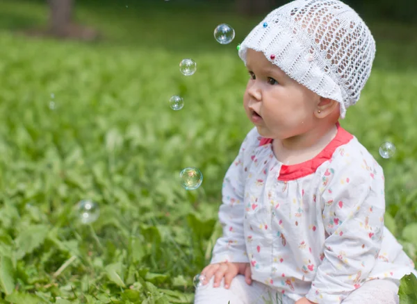 Een klein meisje, zittend op groen gras in het park kijken naar zeep — Stockfoto