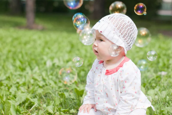 Маленькая девочка сидит на зеленой траве в парке и смотрит на мыло — стоковое фото