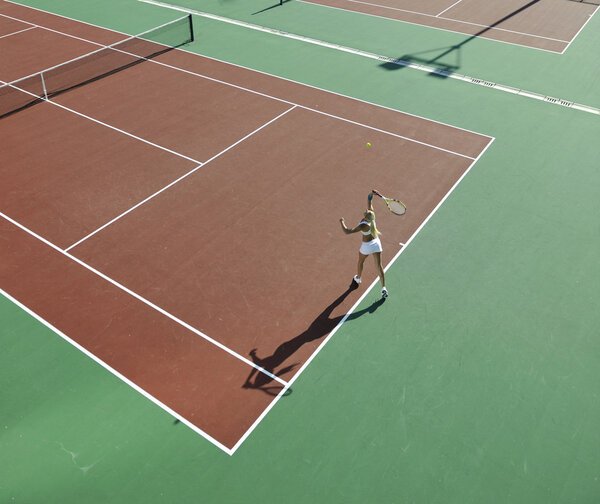 Молодая женщина играет в теннис на открытом воздухе на оранжевом теннисном поле рано утром
