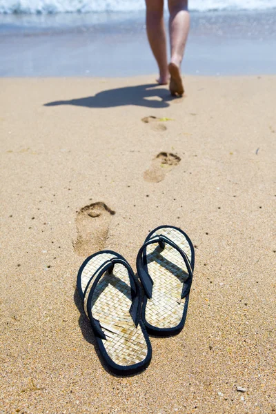 Pantoufles de plage sur le sable et la fille dans l'océan — Photo
