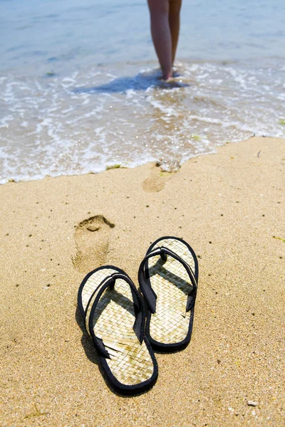 Пляжные тапочки на песке и девушка в океане — стоковое фото