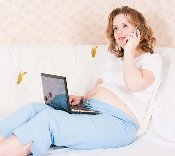 La femme enceinte avec l'ordinateur portable et un téléphone portable sur un canapé — Photo