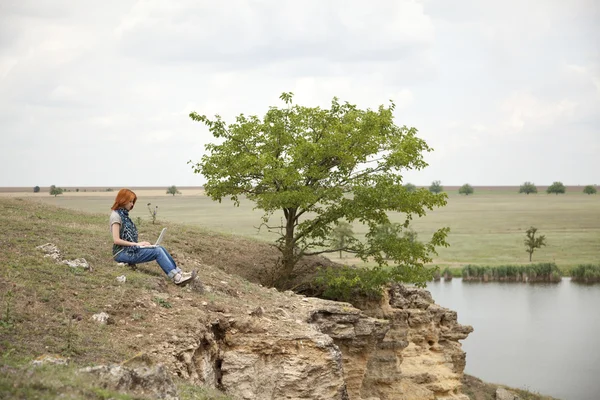 Joven hermosa chica con portátil en la roca cerca del lago y el árbol . —  Fotos de Stock