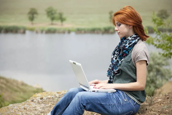 Menina bonita nova com laptop na rocha perto do lago e da árvore . — Fotografia de Stock