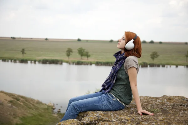 Menina de moda jovem com fones de ouvido na rocha perto do lago . — Fotografia de Stock