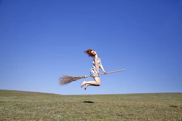Fiatal vörös hajú boszorkány, a zöld fű mező felett repülő seprű — Stock Fotó