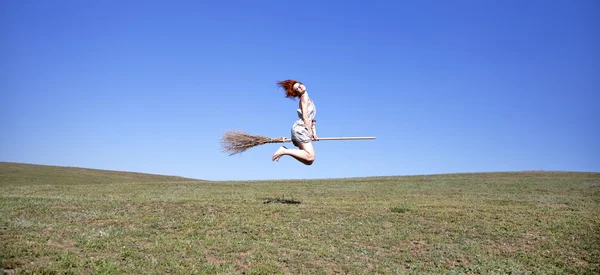 Young red-haired witch on broom flying over green grass field — Stock Photo, Image