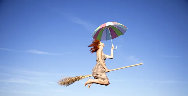 Young red-haired witch on broom flying in the sky with umbrella — Stock Photo, Image