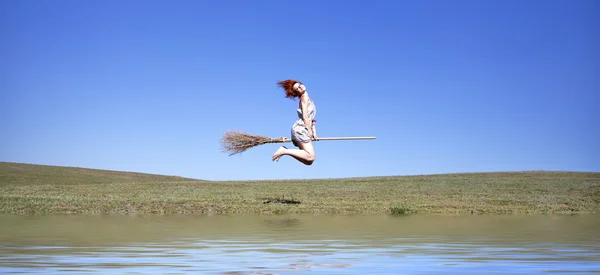 Red-haired witch on broom flying over grass field and water — Stock Photo, Image