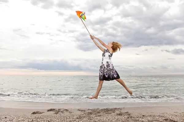 Blond flicka hoppa med vindkraftverk på stranden. — Stockfoto
