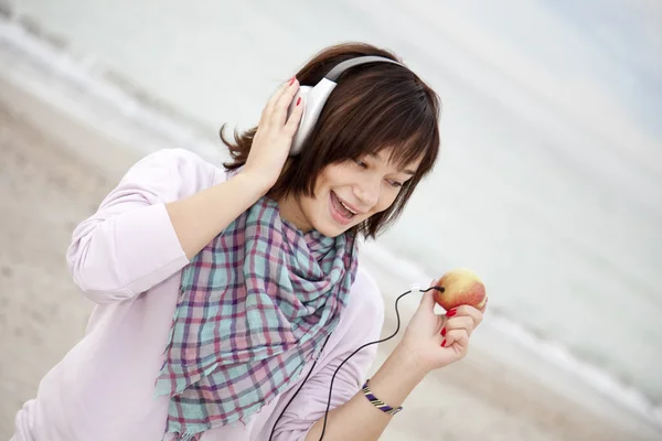 Menina morena jovem com fones de ouvido e maçã na praia . — Fotografia de Stock