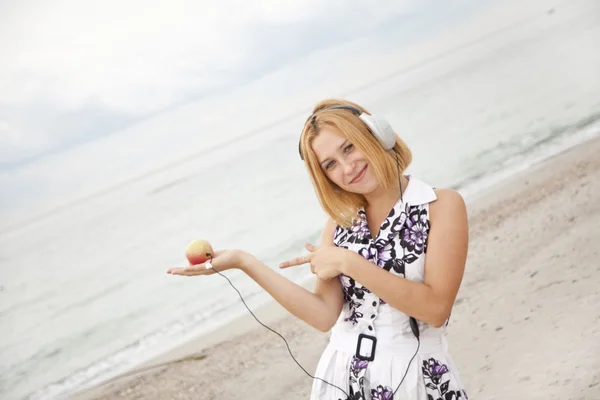 Jonge blonde meisje met hoofdtelefoons en apple op het strand. — Stockfoto