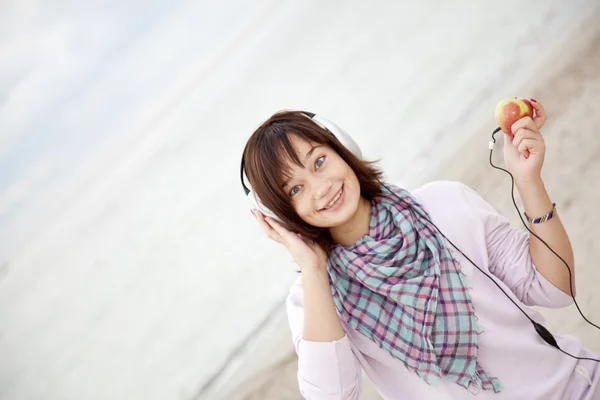 Brunet meisje met hoofdtelefoons en apple op het strand. — Stockfoto
