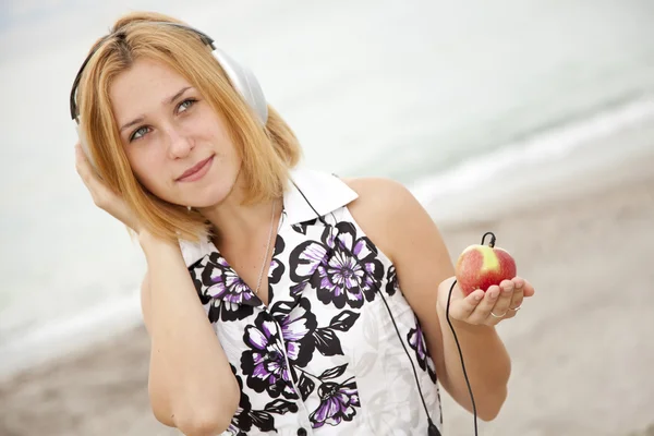 Jonge blonde meisje met hoofdtelefoons en apple op het strand. — Stockfoto