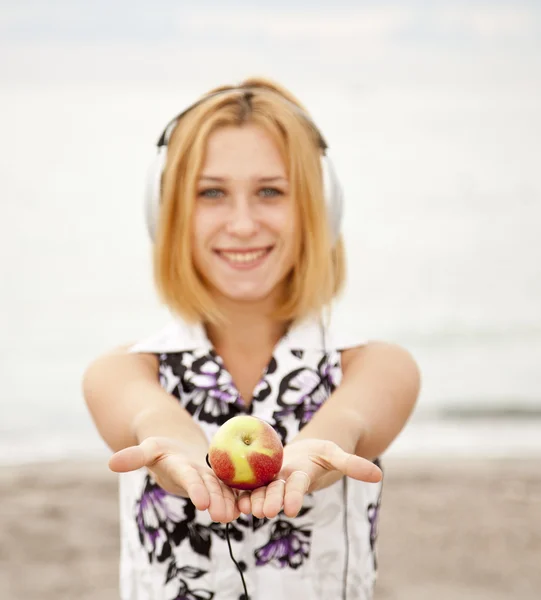 Ung blond tjej med hörlurar och apple på stranden. — Stockfoto