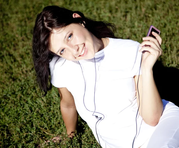 Pretty young brunette girl listening music on green grass in the — Stock Photo, Image